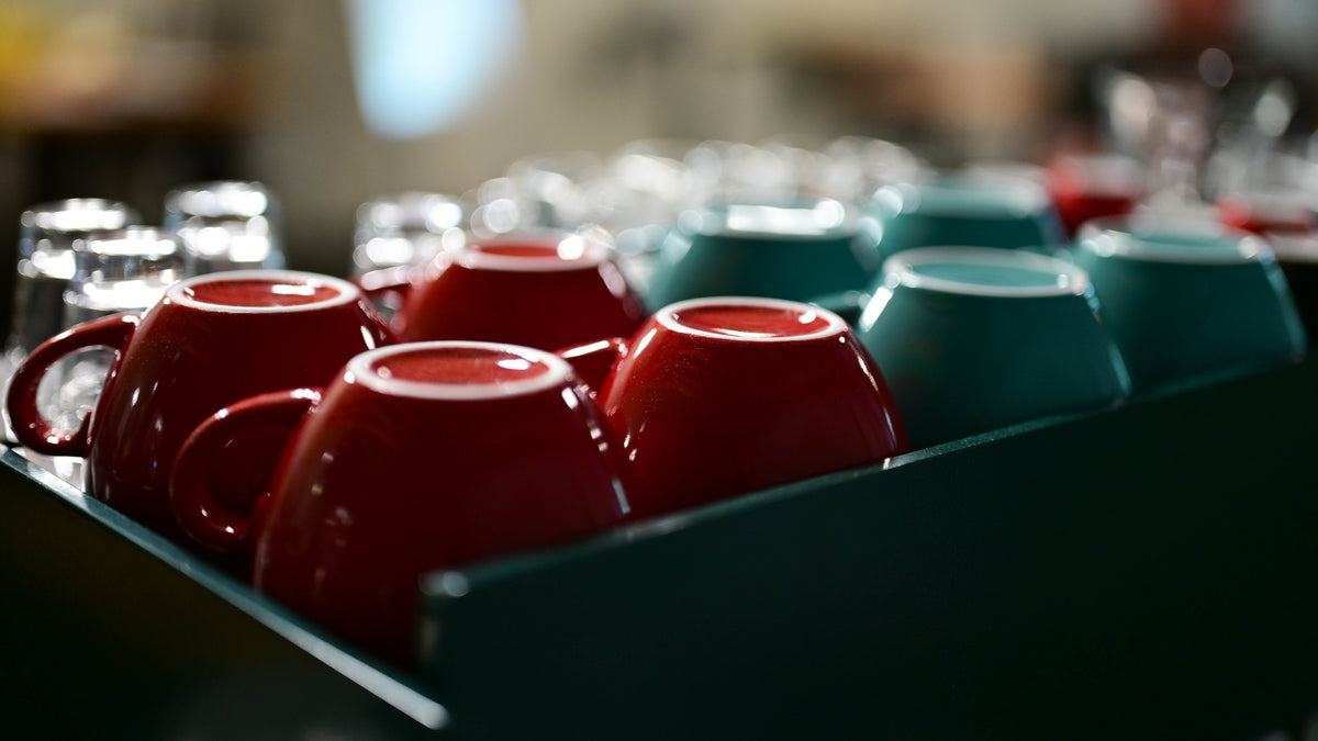 A close-up of glossy ceramic mugs in red and teal arranged in a tray, with a softly blurred background showing more cups. Trail Blaze Coffee Academy.